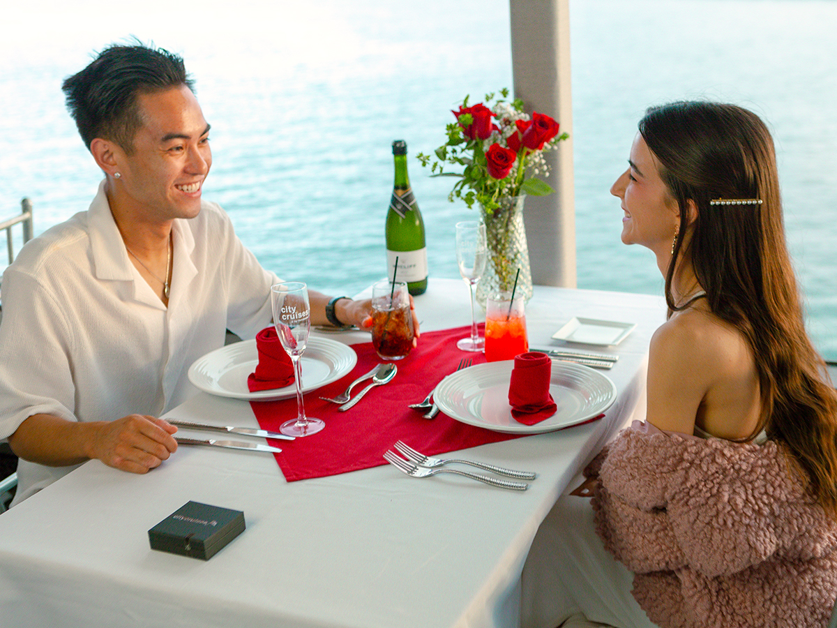 Um casal sentado de frente um para o outro em uma mesa com toalha de mesa branca e um vaso de rosas vermelhas enquanto desfruta de bebidas a bordo de um cruzeiro com jantar.