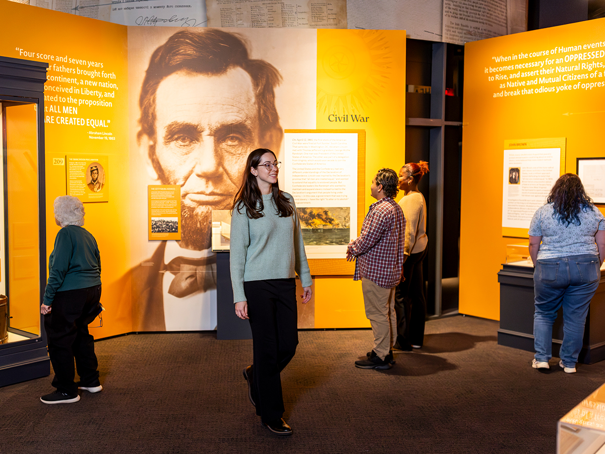 Os visitantes exploram uma nova exposição no Museum of the American Revolution com artefatos, painéis de texto e uma grande imagem de Abraham Lincoln na galeria.