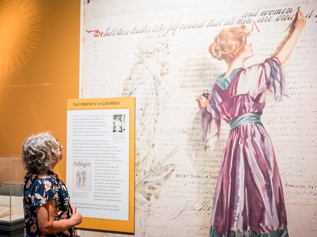 A visitor reads an exhibit panel while looking up at a large mural of a woman writing "and women" into the Declaration of Independence.