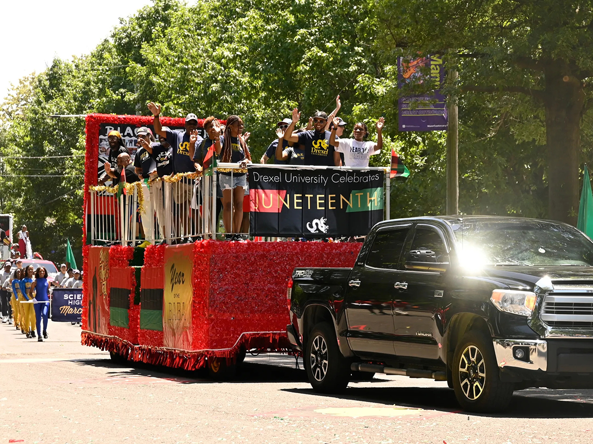Participants wave from a red-decorated parade float as it is pulled down a tree-lined street by a black pick up truck.