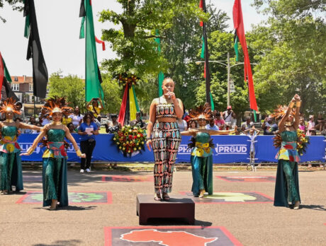 A vocalist stands on a platform while holding a microphone as dancers in shimmering green costumes and feathered headpieces perform behind her at the Juneteenth Parade and Festival.