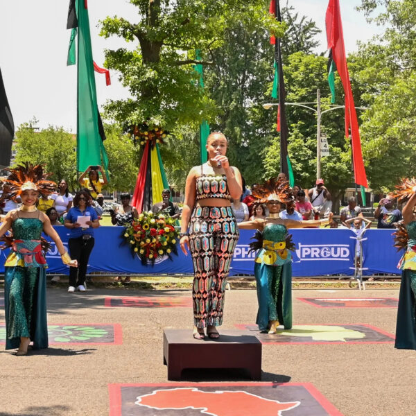 Una cantante se encuentra de pie sobre una plataforma sosteniendo un micrófono, mientras bailarines con brillantes trajes verdes y tocados de plumas actúan detrás de ella en el desfile y festival Juneteenth.