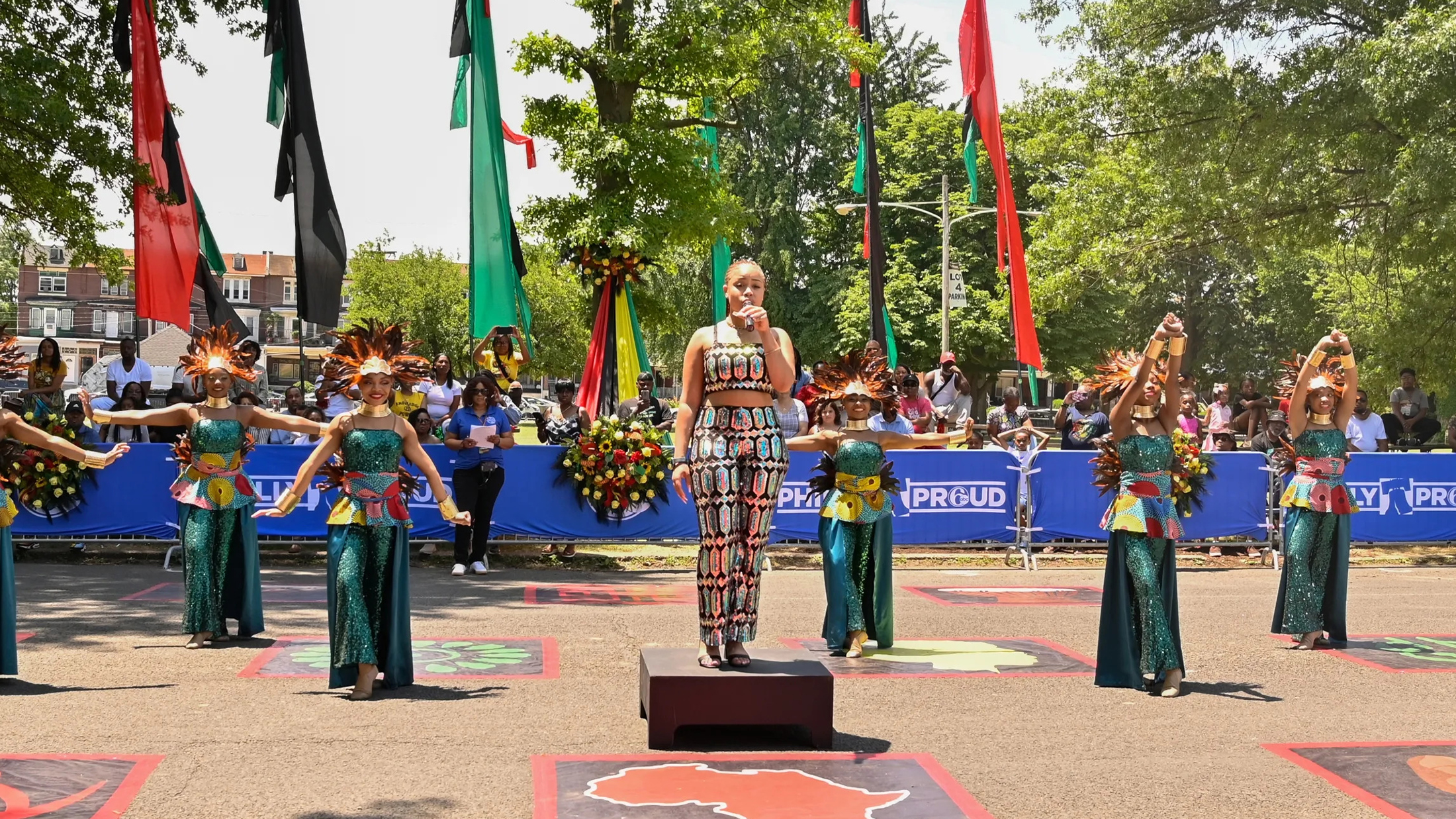 A vocalist stands on a platform while holding a microphone as dancers in shimmering green costumes and feathered headpieces perform behind her at the Juneteenth Parade and Festival.
