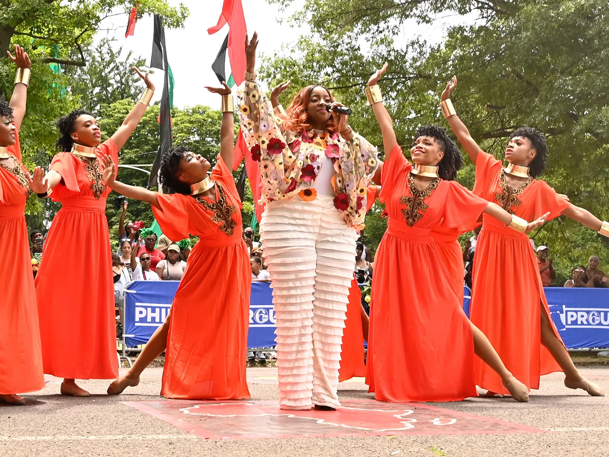 A performer sings into a microphone while surrounded by dancers in flowing orange dresses with gold accessories, their arms raised in unison.