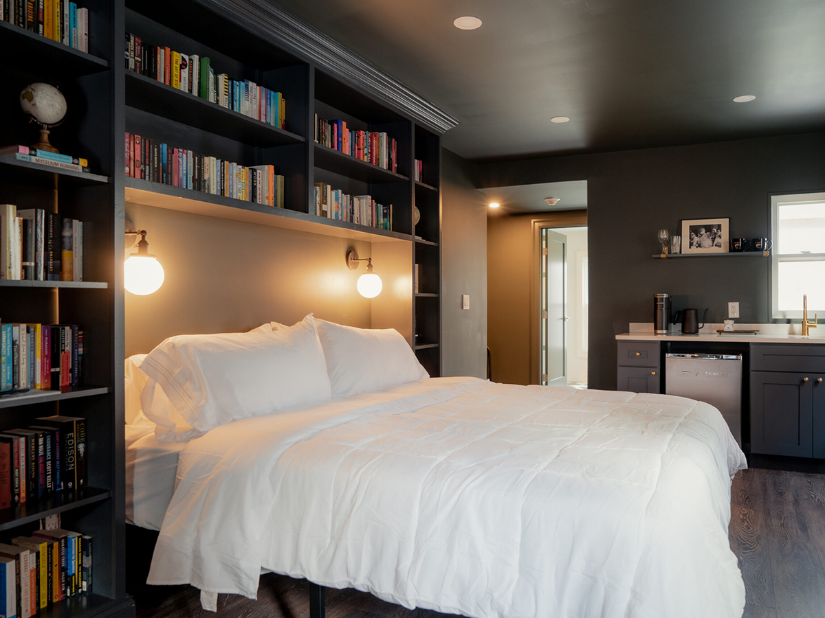 Une chambre moderne de l'hôtel Bookhouse comprend un lit aux draps blancs, encadré par des étagères encastrées, avec un éclairage chaleureux et une kitchenette compacte en arrière-plan.