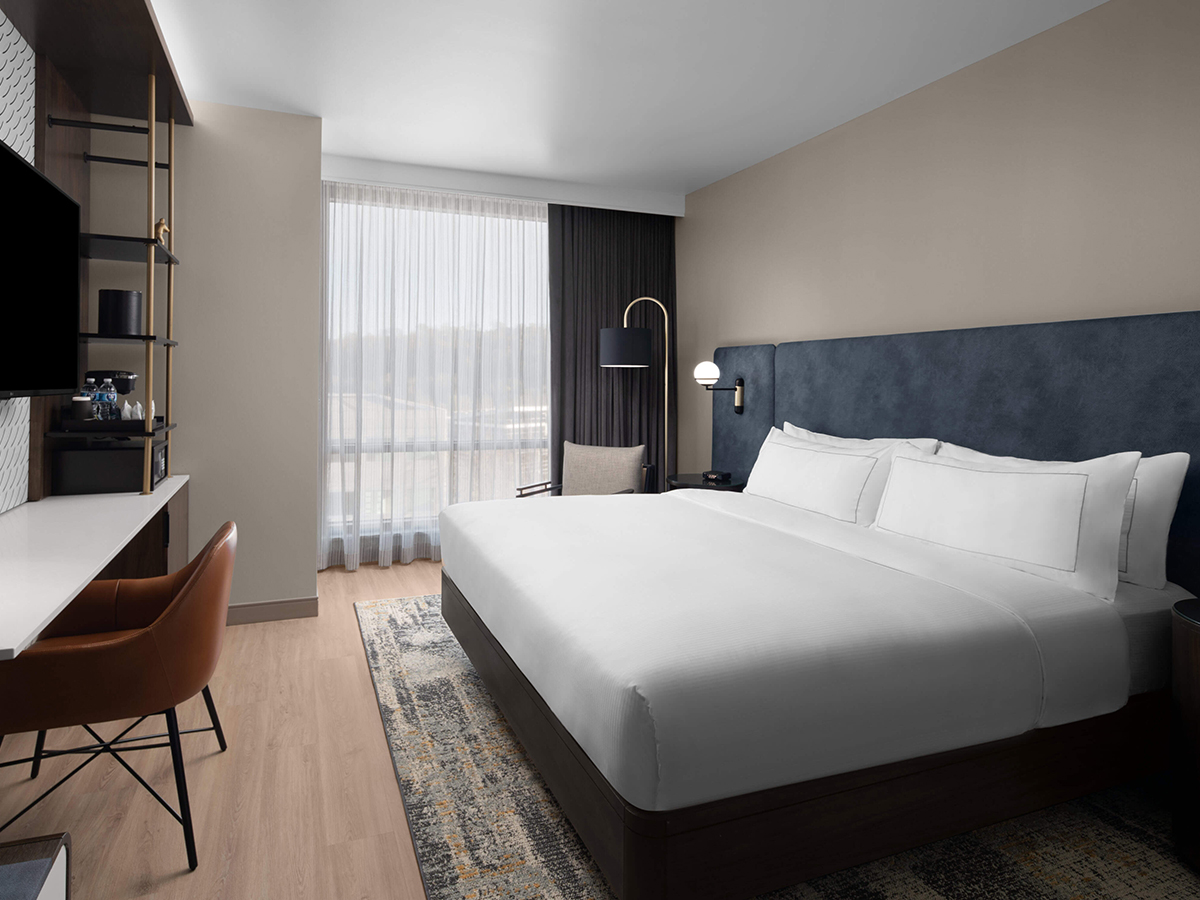 A modern guestroom at Hotel West & Main features a king bed with crisp white linens, a dark blue upholstered headboard, a desk and large windows with sheer curtains.