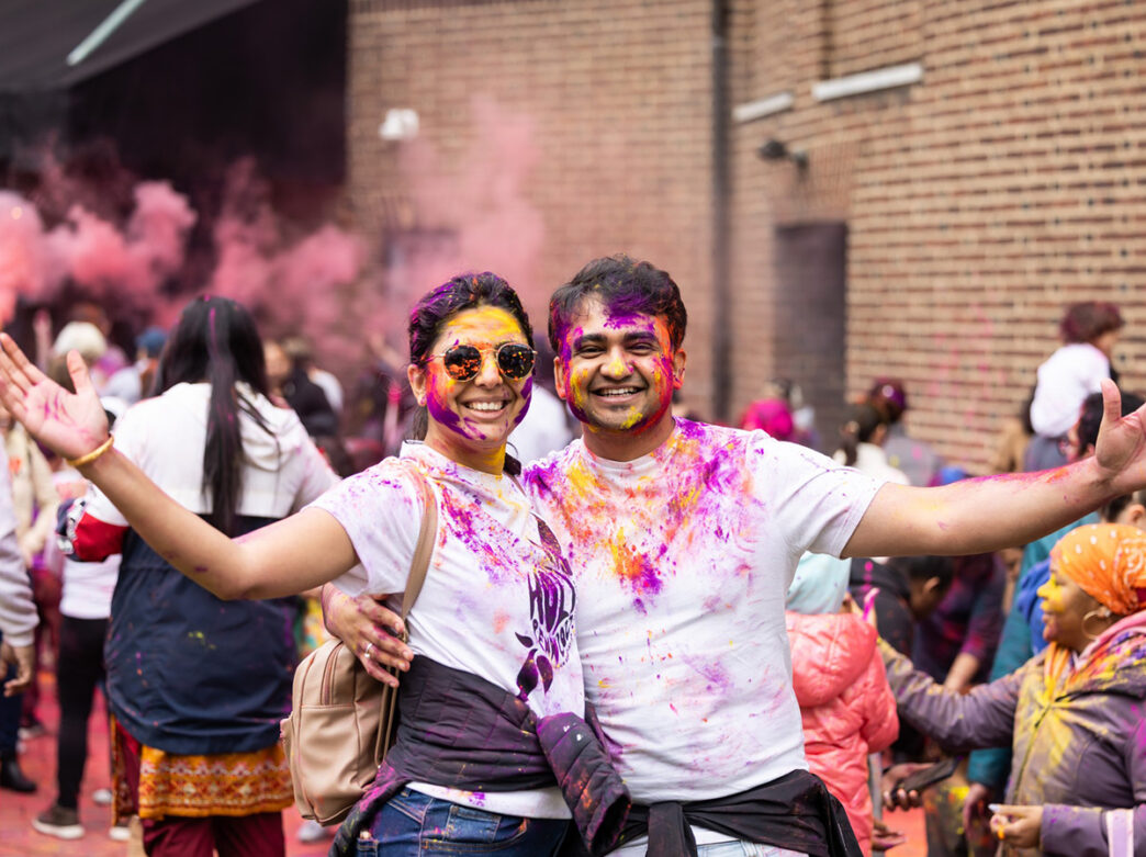 Two smiling people covered in pink, yellow and purple powder pose with arms outstretched during a living Holi celebration at Penn Museum.