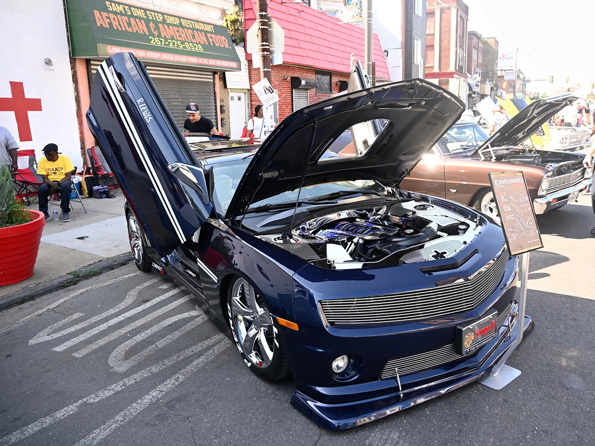 A customized dark blue sports car with its hood and scissor door raised is displayed on a street during the Juneteenth Parade and Festival.