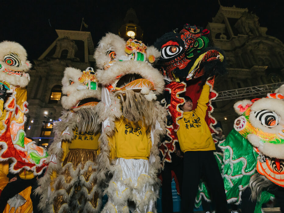 Lion dancers wearing yellow hoodies hold up ornate white, green and red lion costumes while performing at night in front of Philadelphia City Hall.