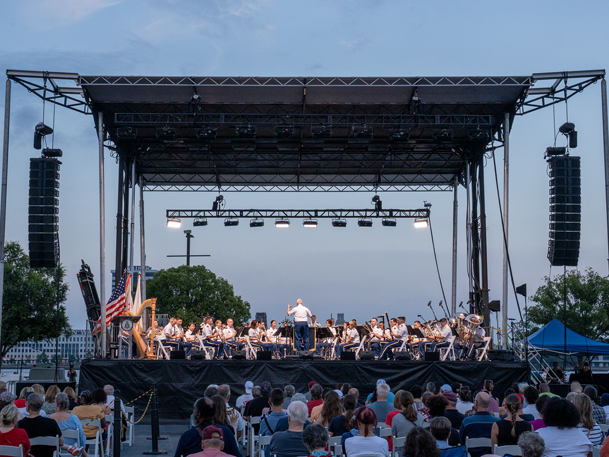 A full orchestra performs on a large outdoor stage at dusk, with a seated audience watching from rows of white folding chairs in an open public space.