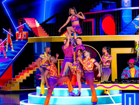 A group of brightly dressed circus performers striking dynamic poses on a colorful, geometric stage set, with dancers lifted overhead and musicians performing in the background under vivid neon lighting.