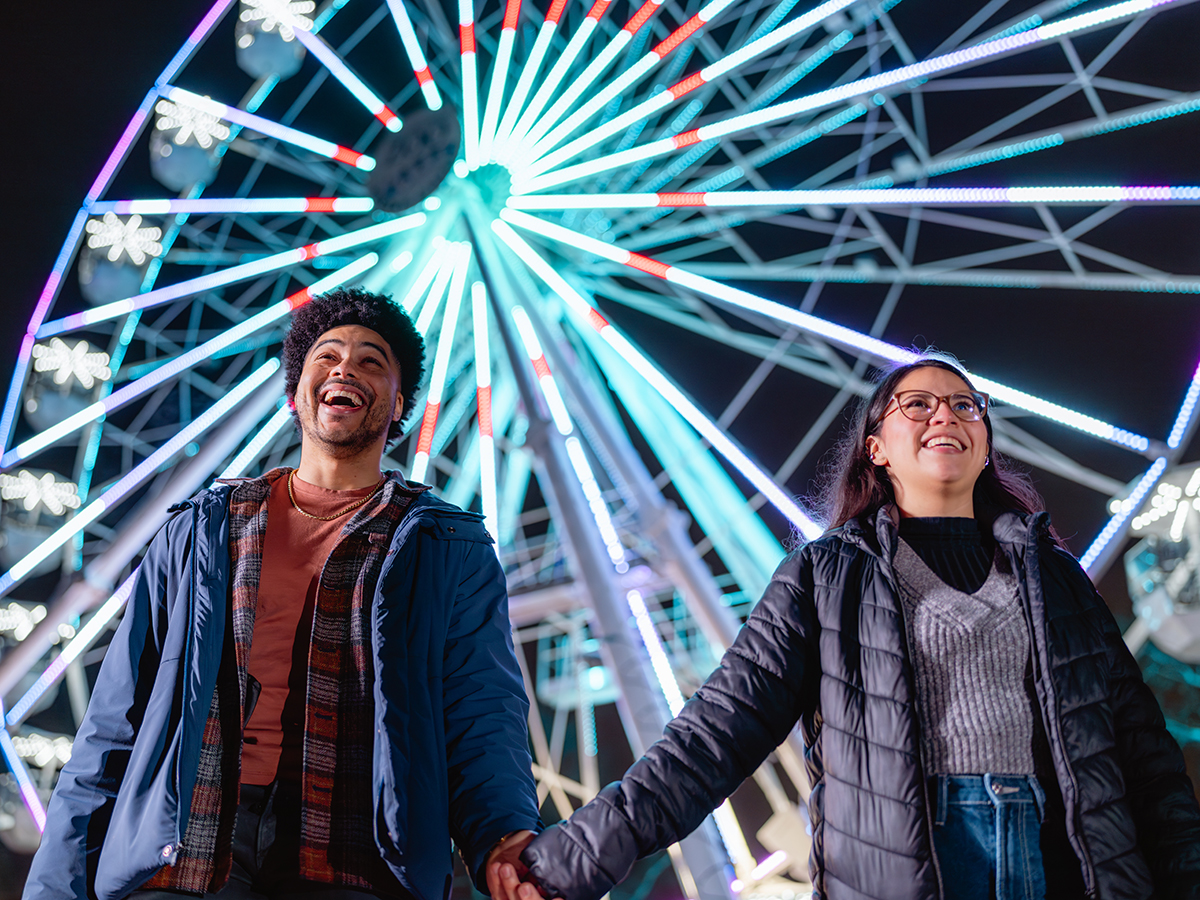 Um casal sorridente dá as mãos enquanto caminha em frente a uma roda-gigante bem iluminada à noite.