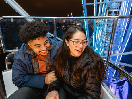 A couple laughs together inside a Ferris wheel gondola at night, surrounded by illuminated metal beams and city lights in the distance.