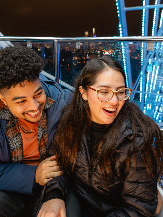 A couple laughs together inside a Ferris wheel gondola at night, surrounded by illuminated metal beams and city lights in the distance.