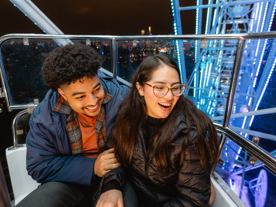 A couple laughs together inside a Ferris wheel gondola at night, surrounded by illuminated metal beams and city lights in the distance.