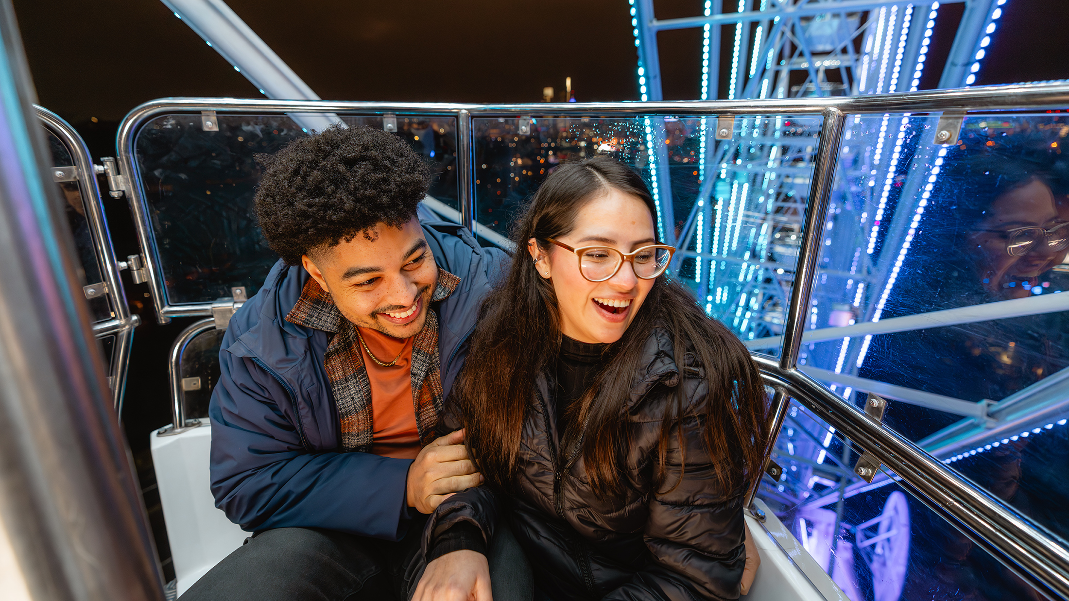 Um casal ri junto em uma gôndola de roda-gigante à noite, cercado por vigas de metal iluminadas e luzes da cidade à distância.
