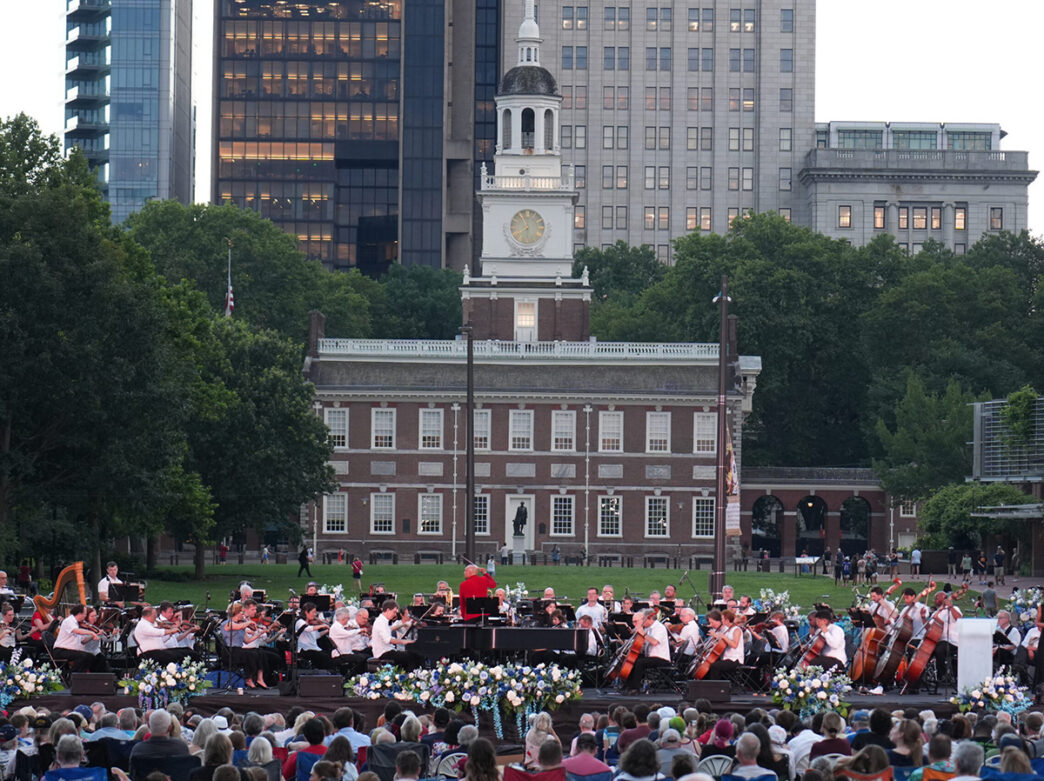 Een orkest treedt buiten op op een podium versierd met bloemen tegen de achtergrond van Independence Hall, met een groot publiek verzameld op het gazon.