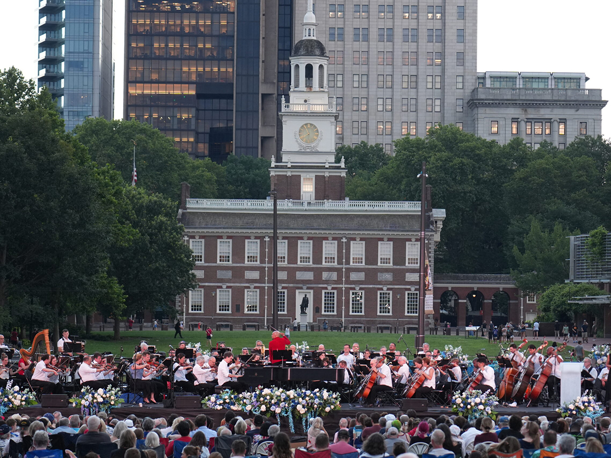 An orchestra performs outdoors on a stage decorated with flowers set against the backdrop of Independence Hall, with a large audience gathered on the lawn.