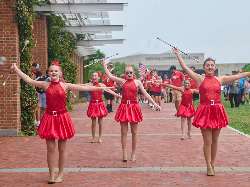 Een rij dansers in rode paillettenjurken speelt met stokken langs een stenen loopbrug tijdens een evenement op 4 juli, met een muziekkorps en toeschouwers achter hen.