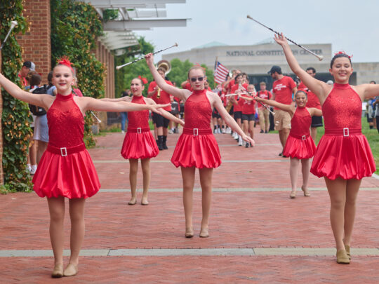 A line of dancers in red sequined dresses perform with batons along a brick walkway during a Fourth of July event, with a marching band and spectators behind them.