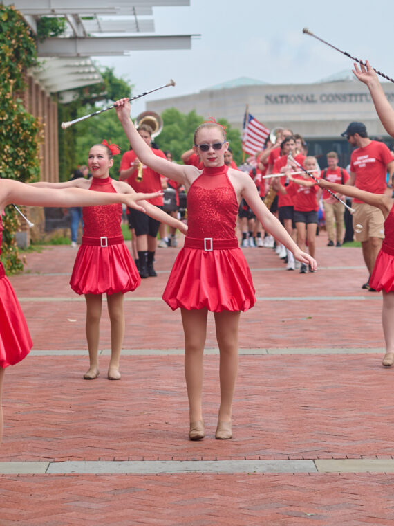 Una fila de bailarinas con vestidos rojos de lentejuelas actúan con bastones a lo largo de una pasarela de ladrillo durante un evento del 4 de julio, con una banda de música y espectadores detrás de ellas.