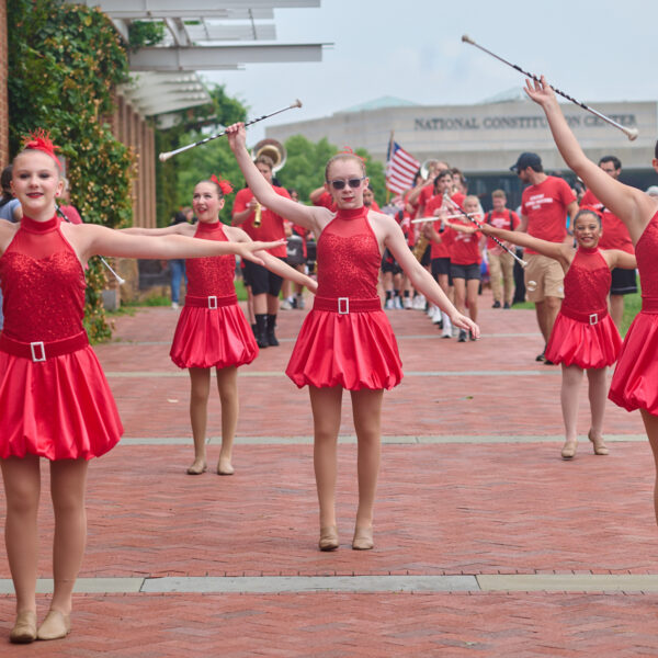 A line of dancers in red sequined dresses perform with batons along a brick walkway during a Fourth of July event, with a marching band and spectators behind them.