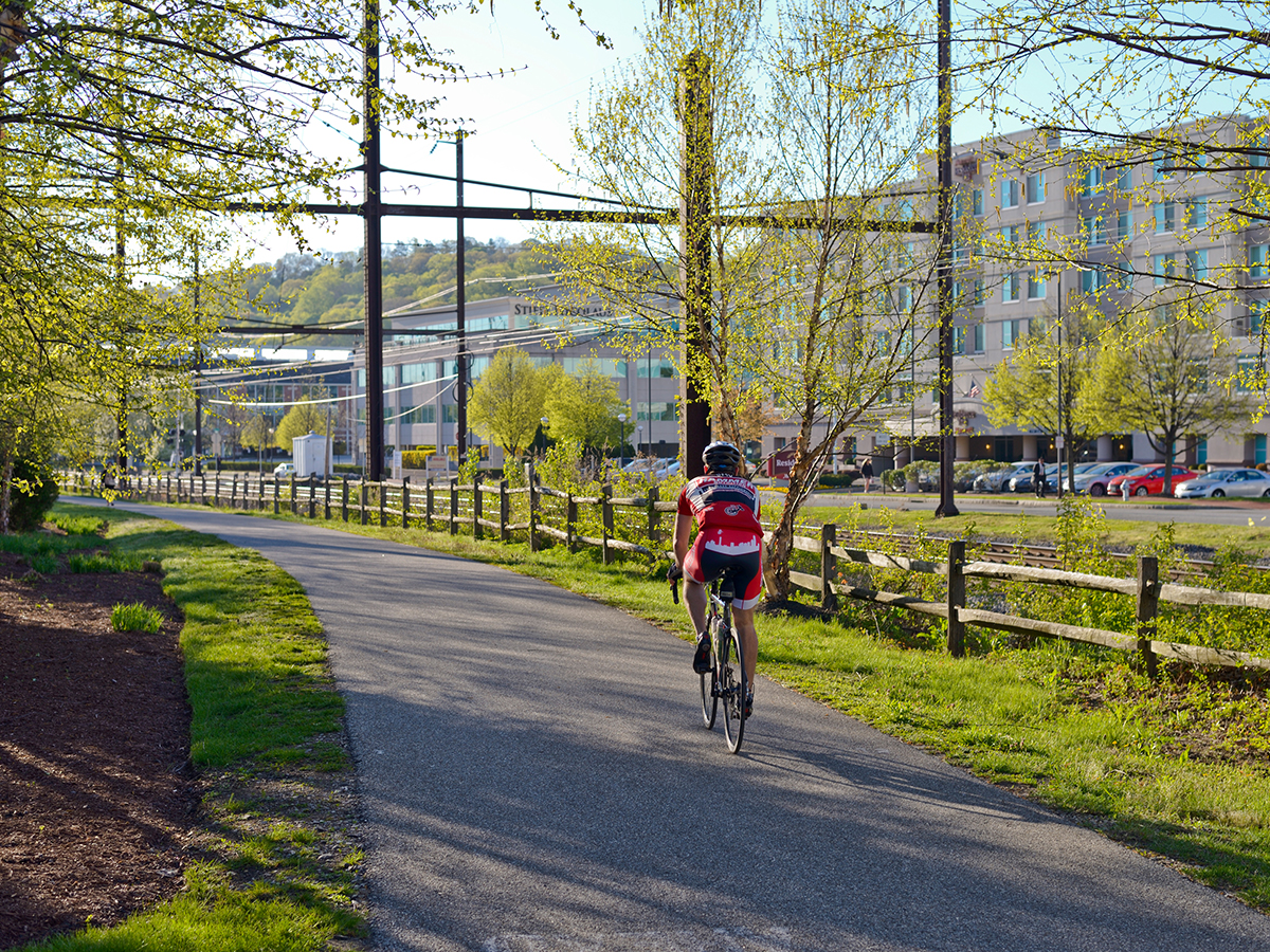 A cyclist rides along a tree-lined paved trail on a sunny day, with office buildings and spring greenery in the background.