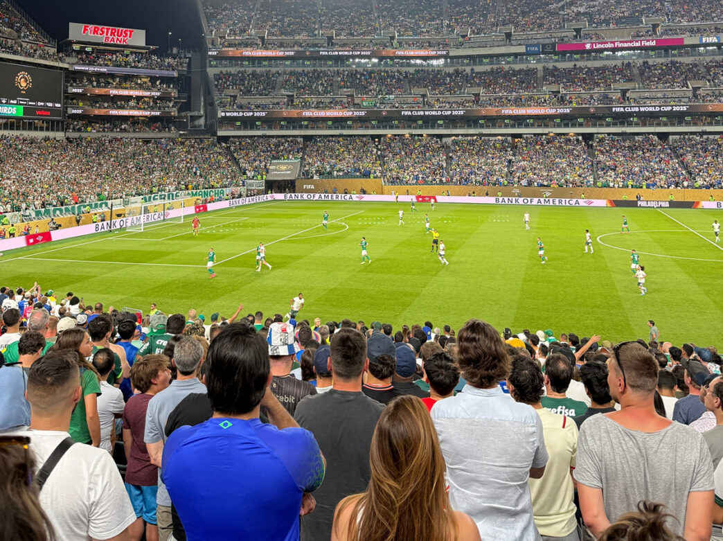 Multitud de espectadores que ven un partido de fútbol en un gran estadio al aire libre. Los jugadores, con camisetas de colores vivos, corren por el césped.
