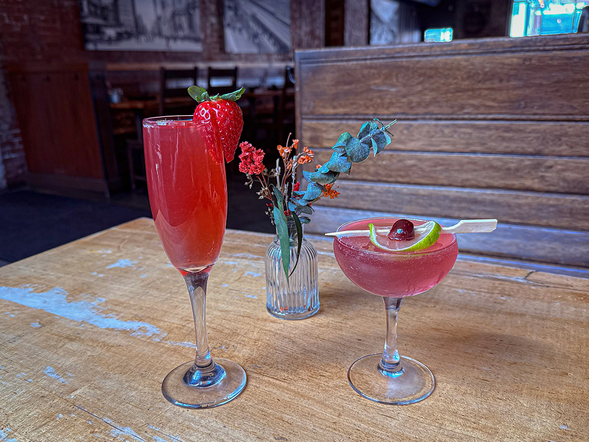 Dois coquetéis cor-de-rosa estão sobre uma mesa de madeira rústica, um em uma taça de champanhe decorada com um morango e o outro em uma taça com uma cereja e casca de limão.