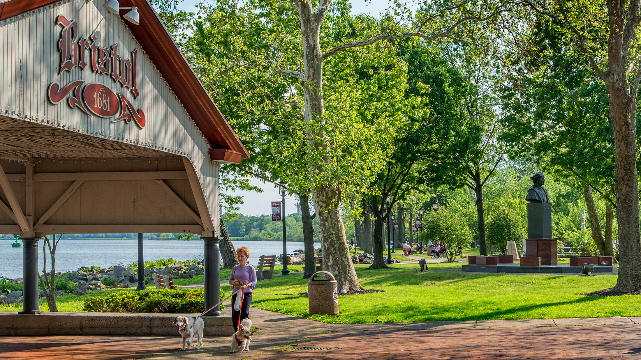 A woman walks her two dogs along the waterfront at Bristol Waterfront Park. The white and red pavilion is boarded by lush green trees.