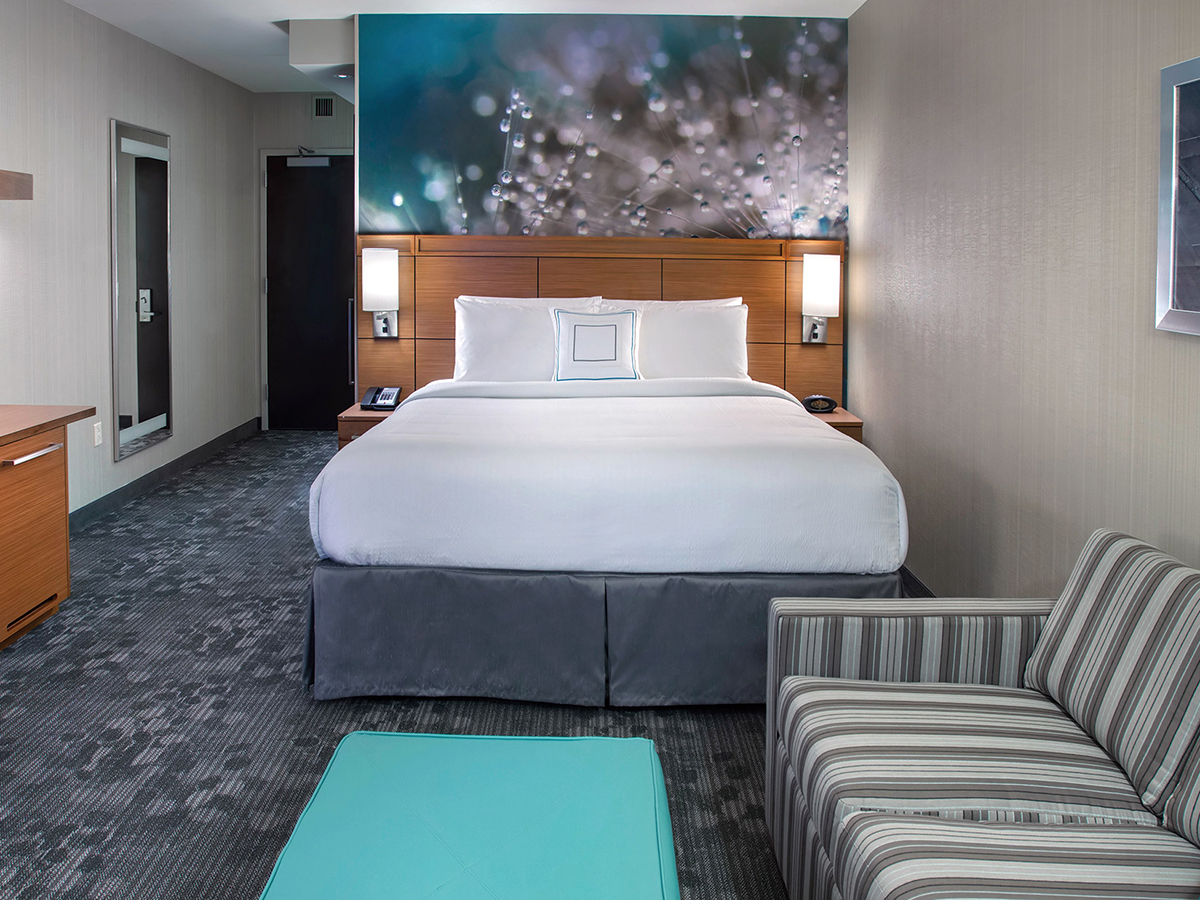 Une chambre d'hôtel à l'intérieur du Courtyard South Philadelphia at the Navy Yard avec un lit king-size blanc et gris, un canapé rayé, une petite table bleu sarcelle et une télévision sur une commode.