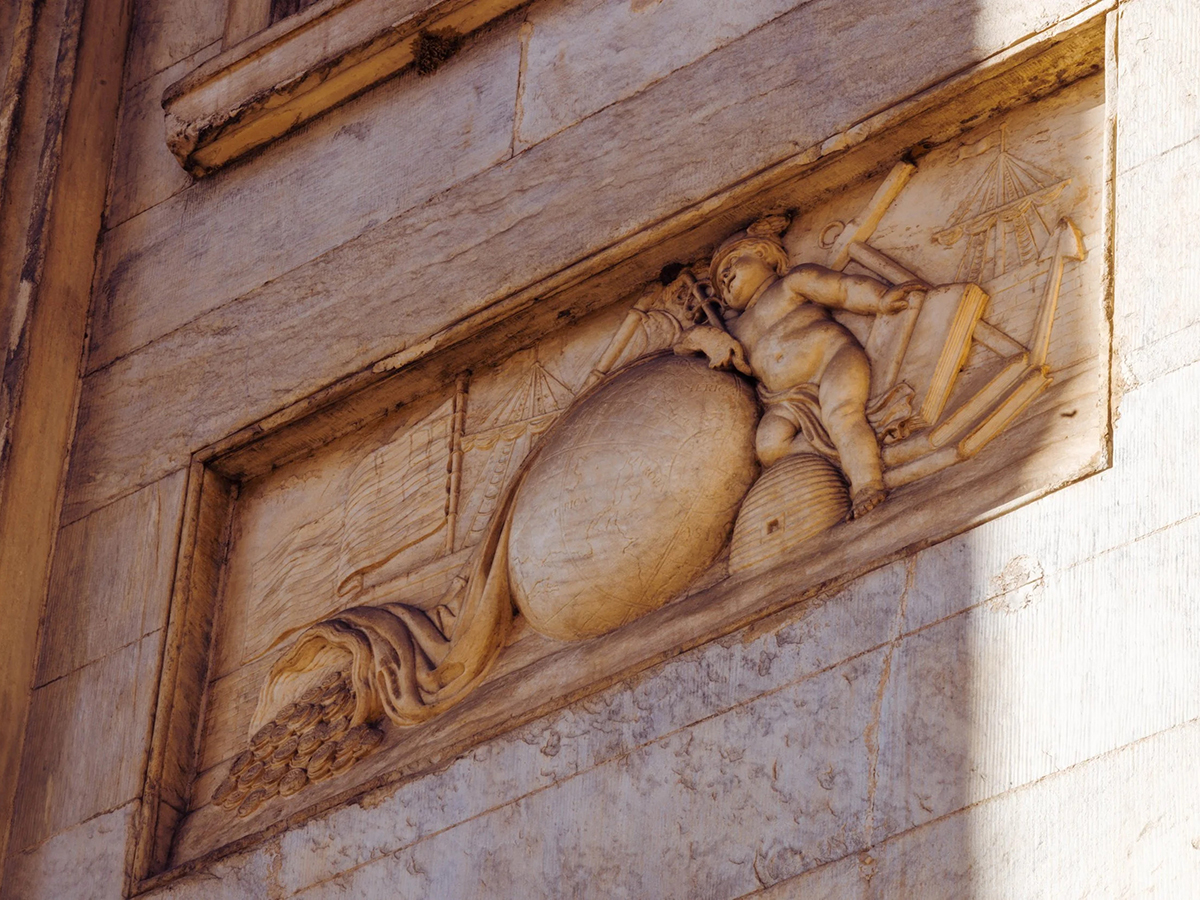 A carved stone relief on the First Bank of the United States exterior depicting allegorical figures alongside a globe, ship and cornucopia.