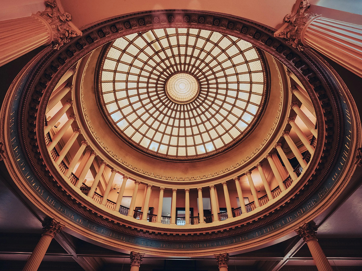 Looking upward to a dramatic circular rotunda topped by a patterned glass dome and surrounded by ring of classical columns.