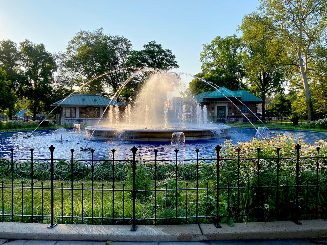 Water sprays from the circular fountain at Franklin Square as sunlight glows through the mist, with flowers, trees and park buildings surrounding the fountain.