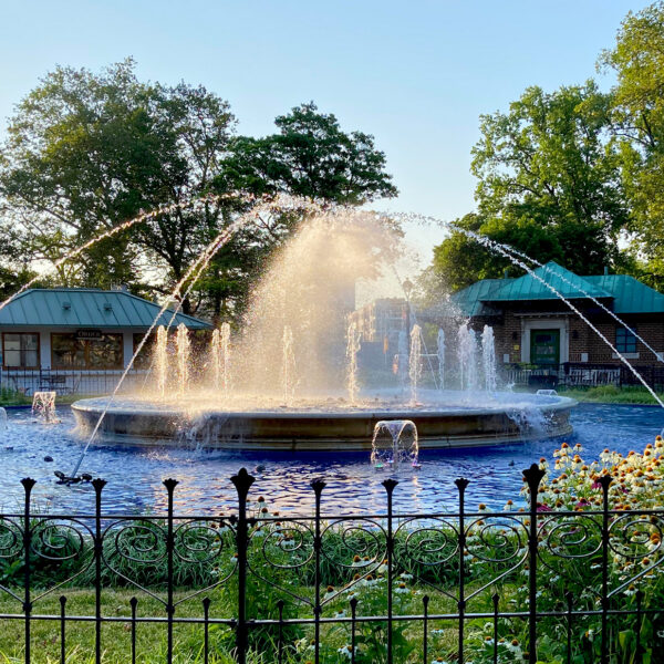 Water sprays from the circular fountain at Franklin Square as sunlight glows through the mist, with flowers, trees and park buildings surrounding the fountain.