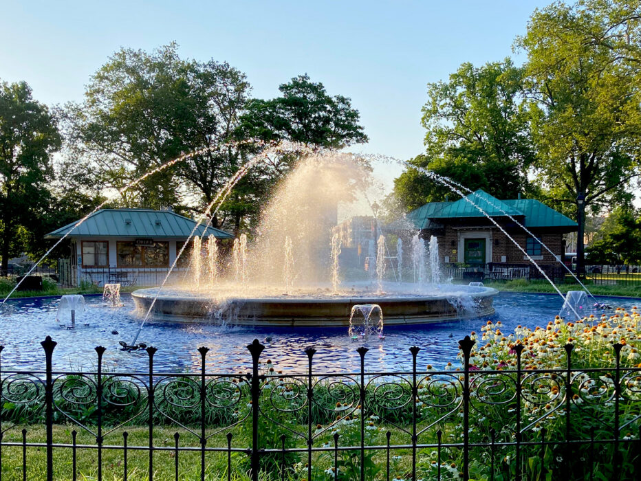El agua brota de la fuente circular en Franklin Square mientras la luz del sol brilla a través de la niebla, con flores, árboles y edificios del parque rodeando la fuente.
