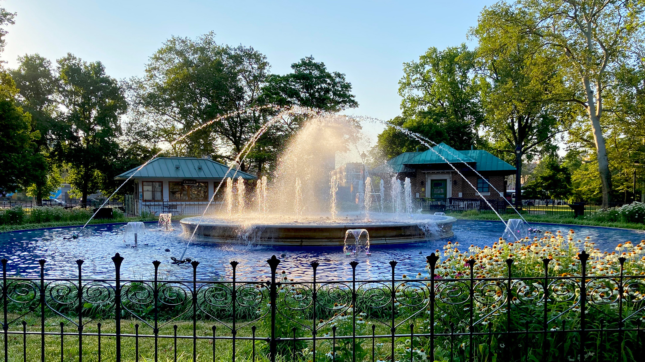 Water spuit uit de ronde fontein op Franklin Square terwijl zonlicht door de mist schijnt, met bloemen, bomen en parkgebouwen die de fontein omringen.