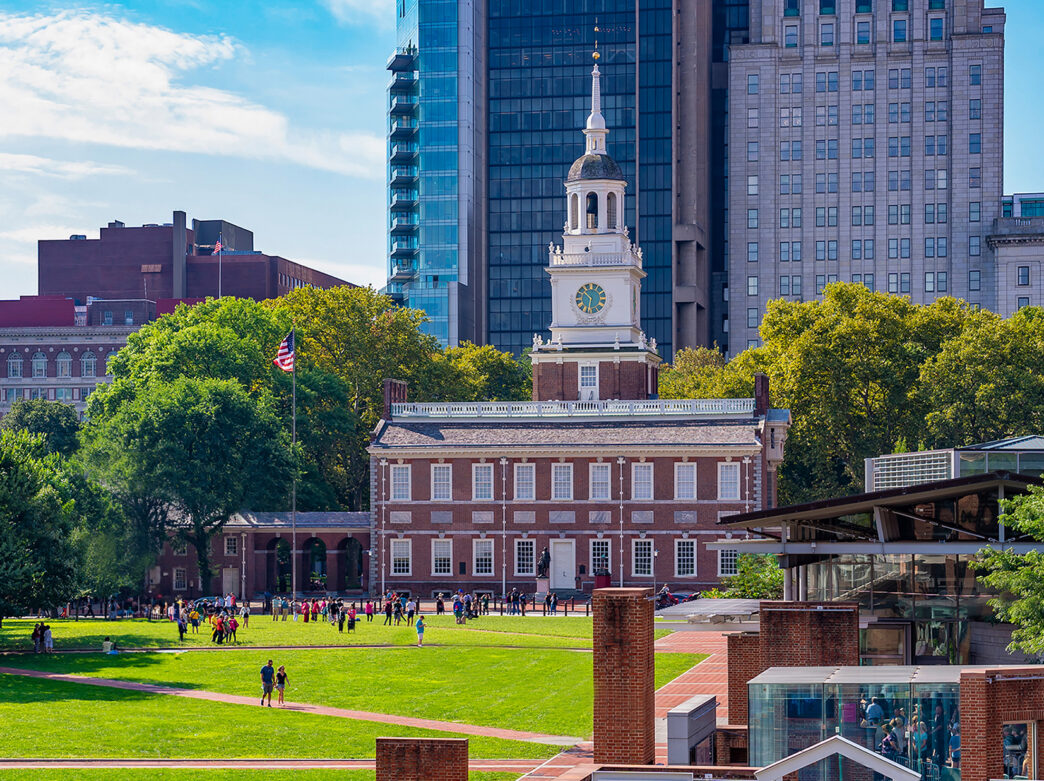 Visitors stroll across the green lawns of Independence Mall, with Independence Hall's iconic clock tower rising above the historic building and the city skyline behind it.