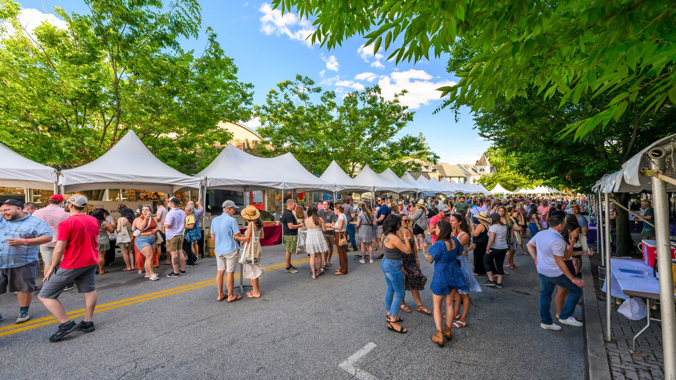 A large crowd of people congregate in front of a row white vendor tents.