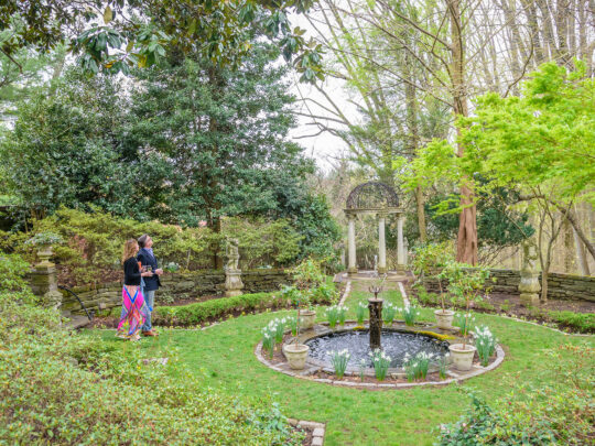 Un couple élégamment vêtu admire le feuillage du début du printemps à Meadowbrook Farm. On aperçoit une fontaine et un belvédère au centre de la photo.