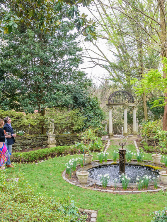 Un couple élégamment vêtu admire le feuillage du début du printemps à Meadowbrook Farm. On aperçoit une fontaine et un belvédère au centre de la photo.