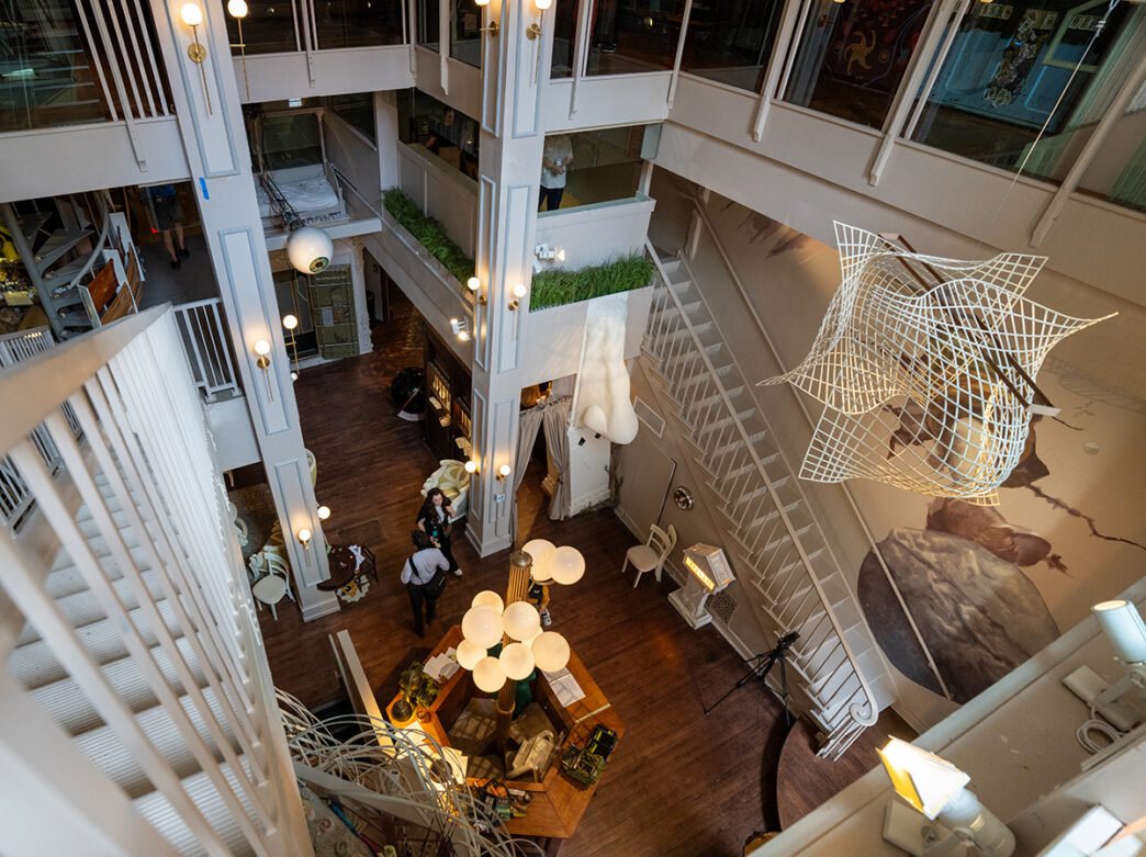 An overhead view of the airy lobby at the Ministry of Awe shows white staircases, sculptural installations and visitors exploring the multilevel space.