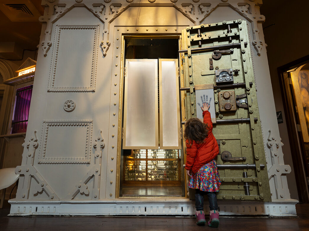 A child reaches up to touch a handprint on the inside of a massive historic vault door at the Ministry of Awe.