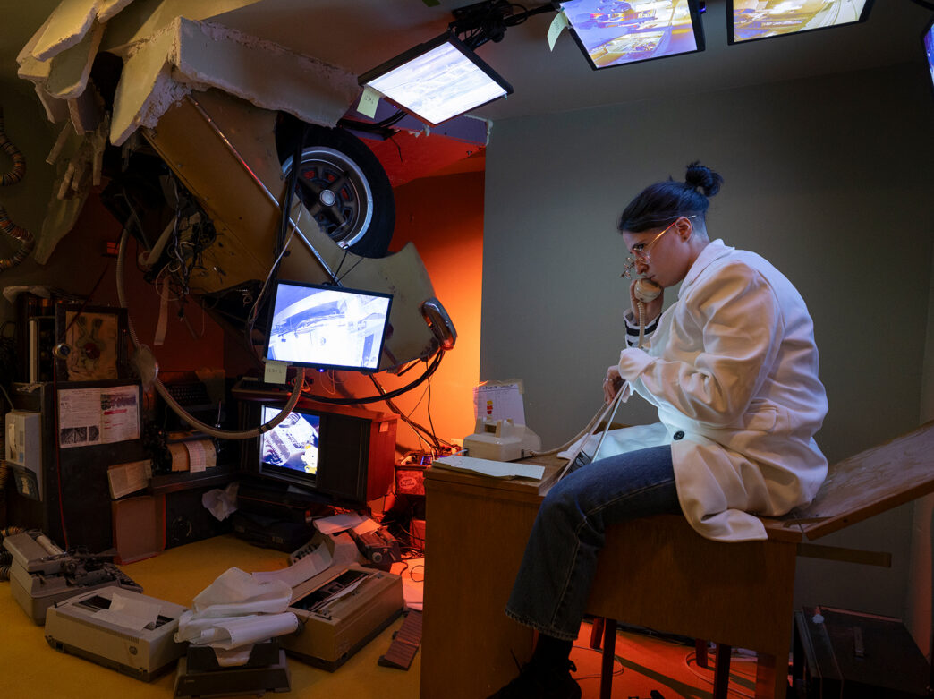 An actor in a lab coat works at a cluttered control station surrounded by monitors and machines in an interactive room at the Ministry of Awe.