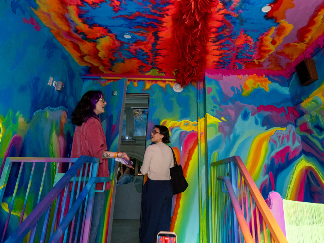 Two visitors walk up a staircase surrounded by vibrant, rainbow-colored murals and textured ceiling at the Ministry of Awe.