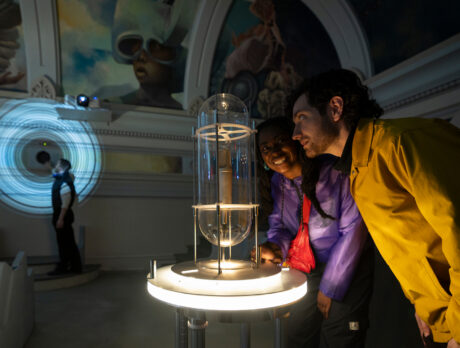 Two visitors lean in to examine a glowing interactive installation inside a dimly lit gallery space at the Ministry of Awe.