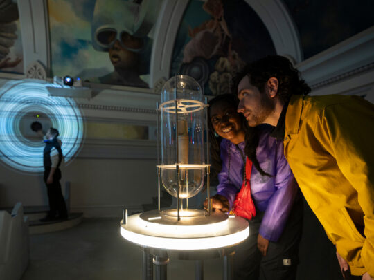 Dos visitantes se inclinan para examinar una instalación interactiva luminosa dentro de una sala de exposiciones con poca luz en el Ministry of Awe.