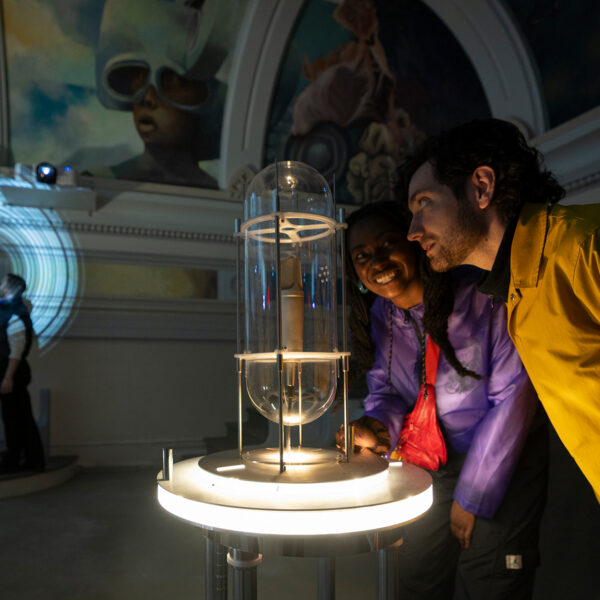 Two visitors lean in to examine a glowing interactive installation inside a dimly lit gallery space at the Ministry of Awe.