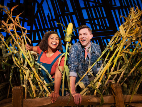 Two performers smile and lean over a wooden fence surrounded by tall cornstalks on a barn-themed stage set from the musical Shucked.