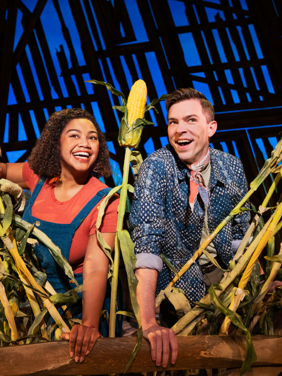 Two performers smile and lean over a wooden fence surrounded by tall cornstalks on a barn-themed stage set from the musical Shucked.